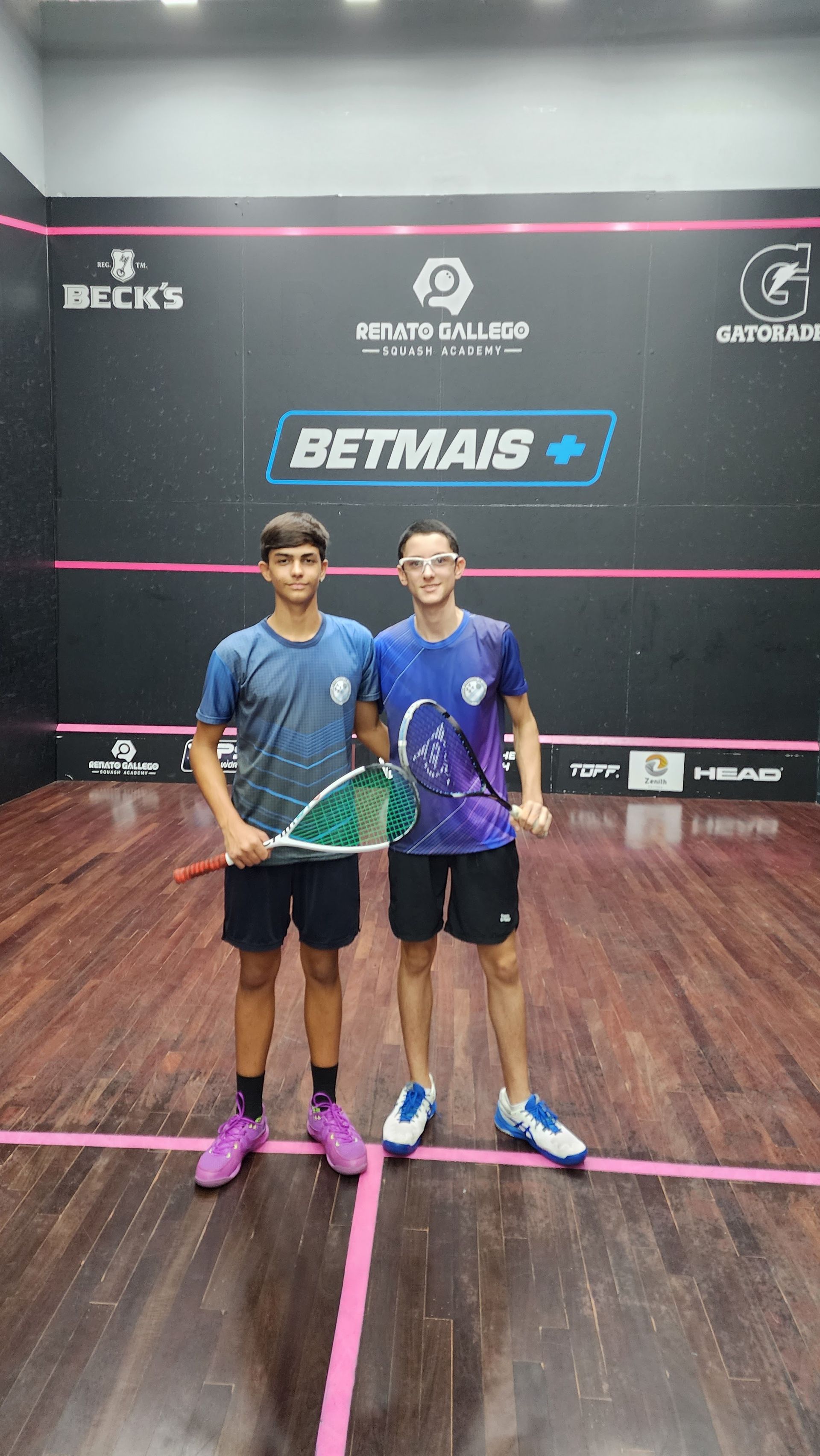Dois jovens estão de pé, um ao lado do outro, em uma quadra de squash.