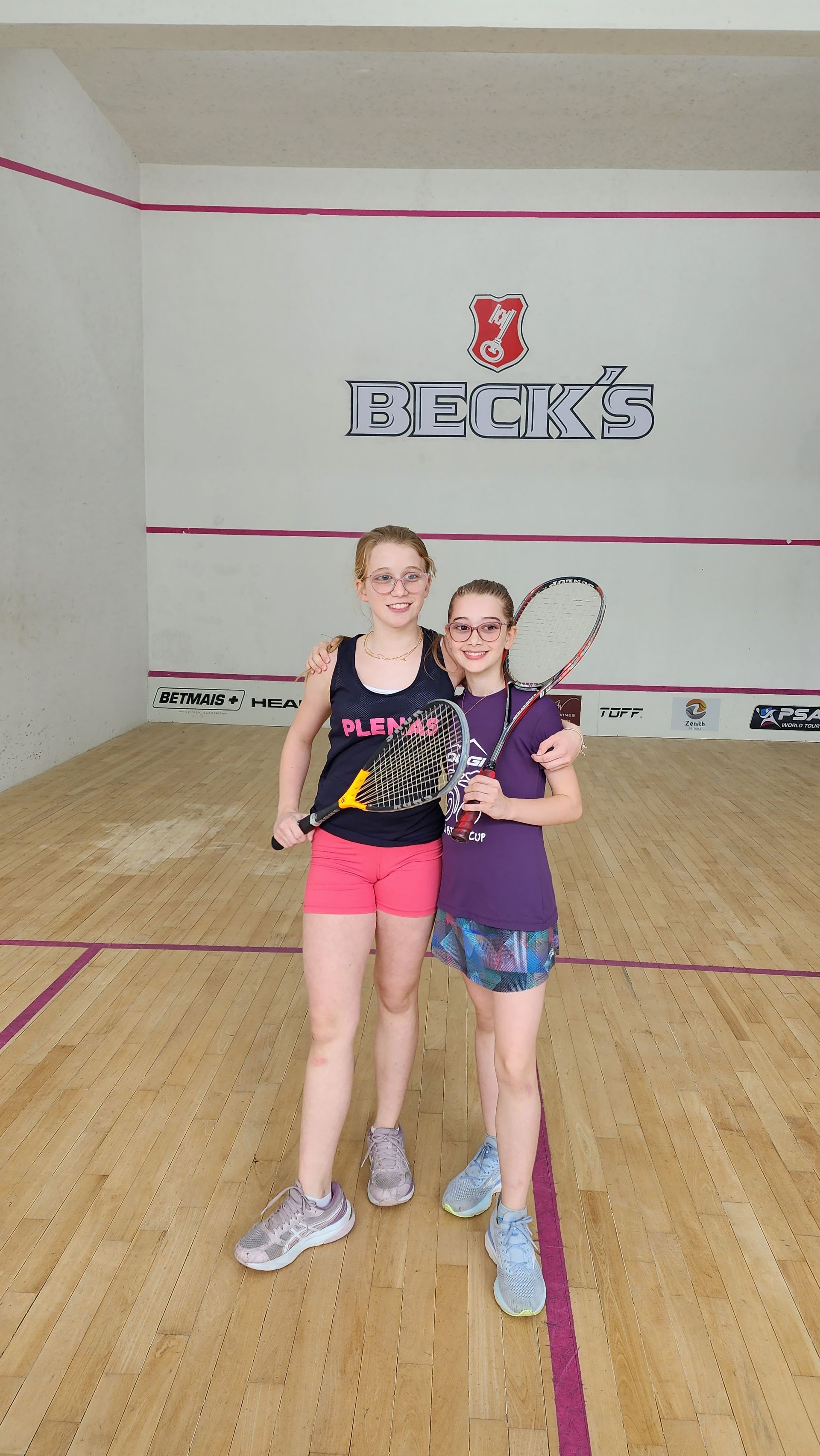 Duas jovens estão posando para uma foto em uma quadra de squash.