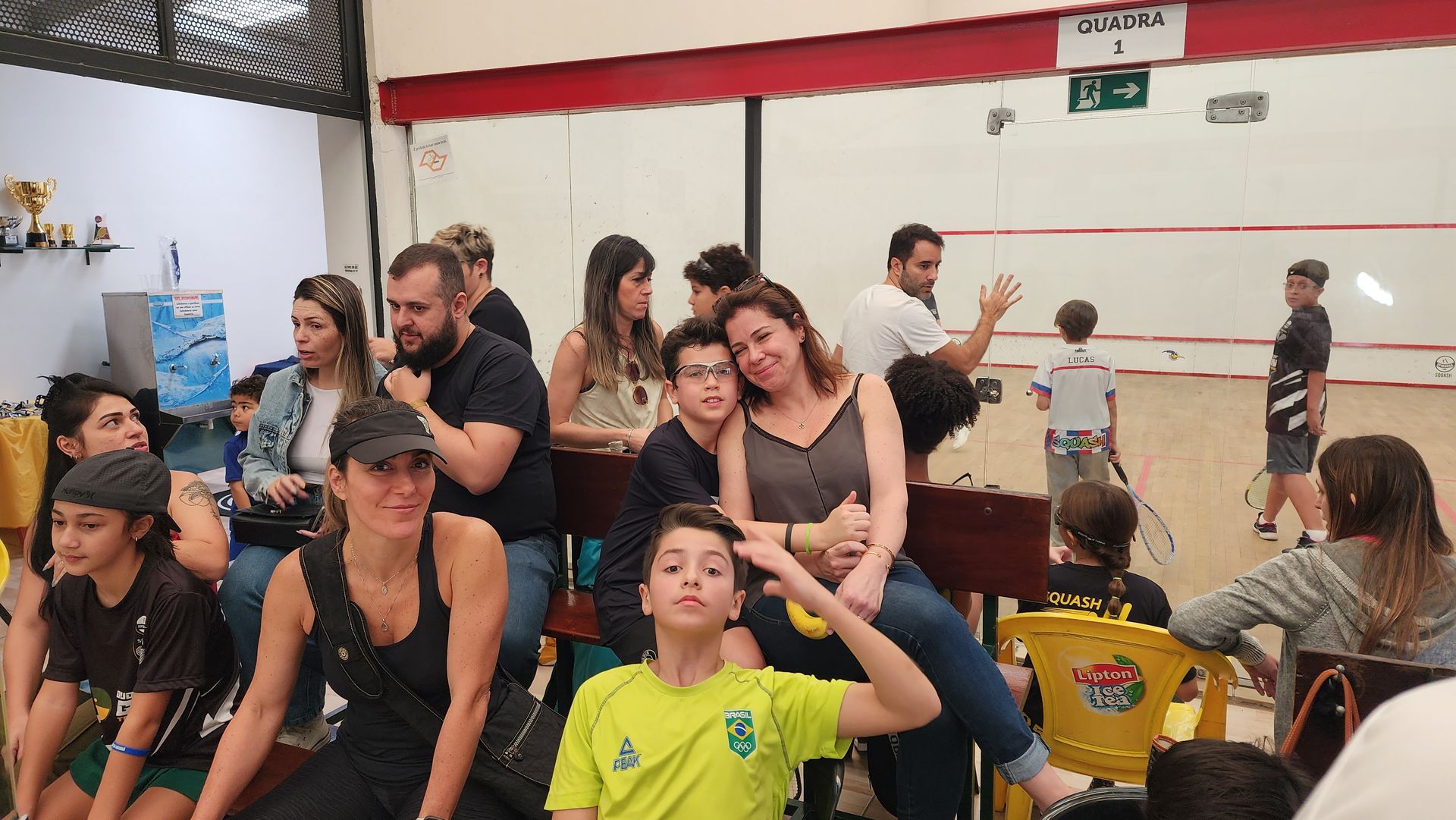 Um grupo de pessoas está sentado em cadeiras em frente a uma quadra de squash.