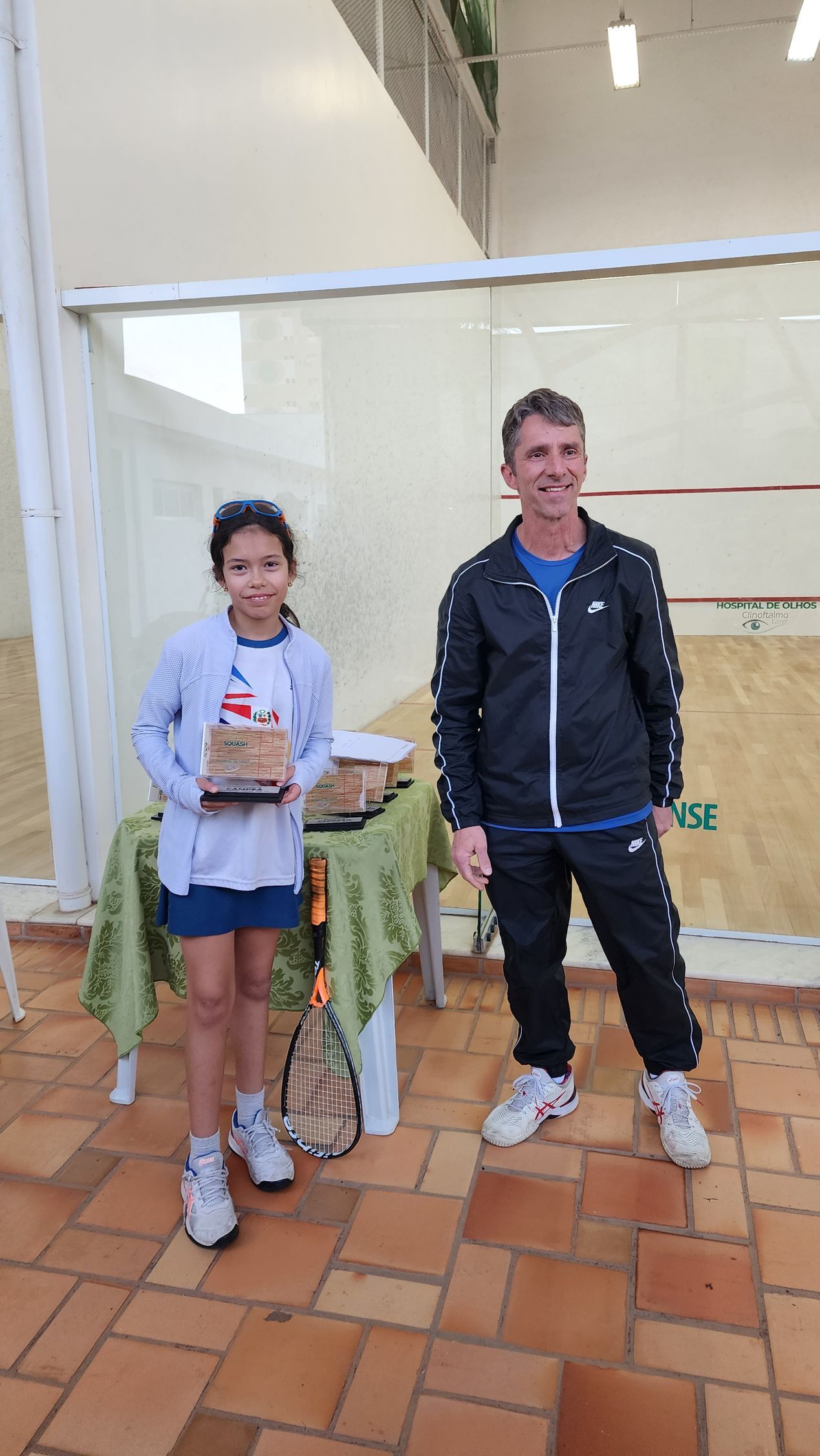 Um homem e uma mulher estão de pé, um ao lado do outro, em frente a uma quadra de squash.