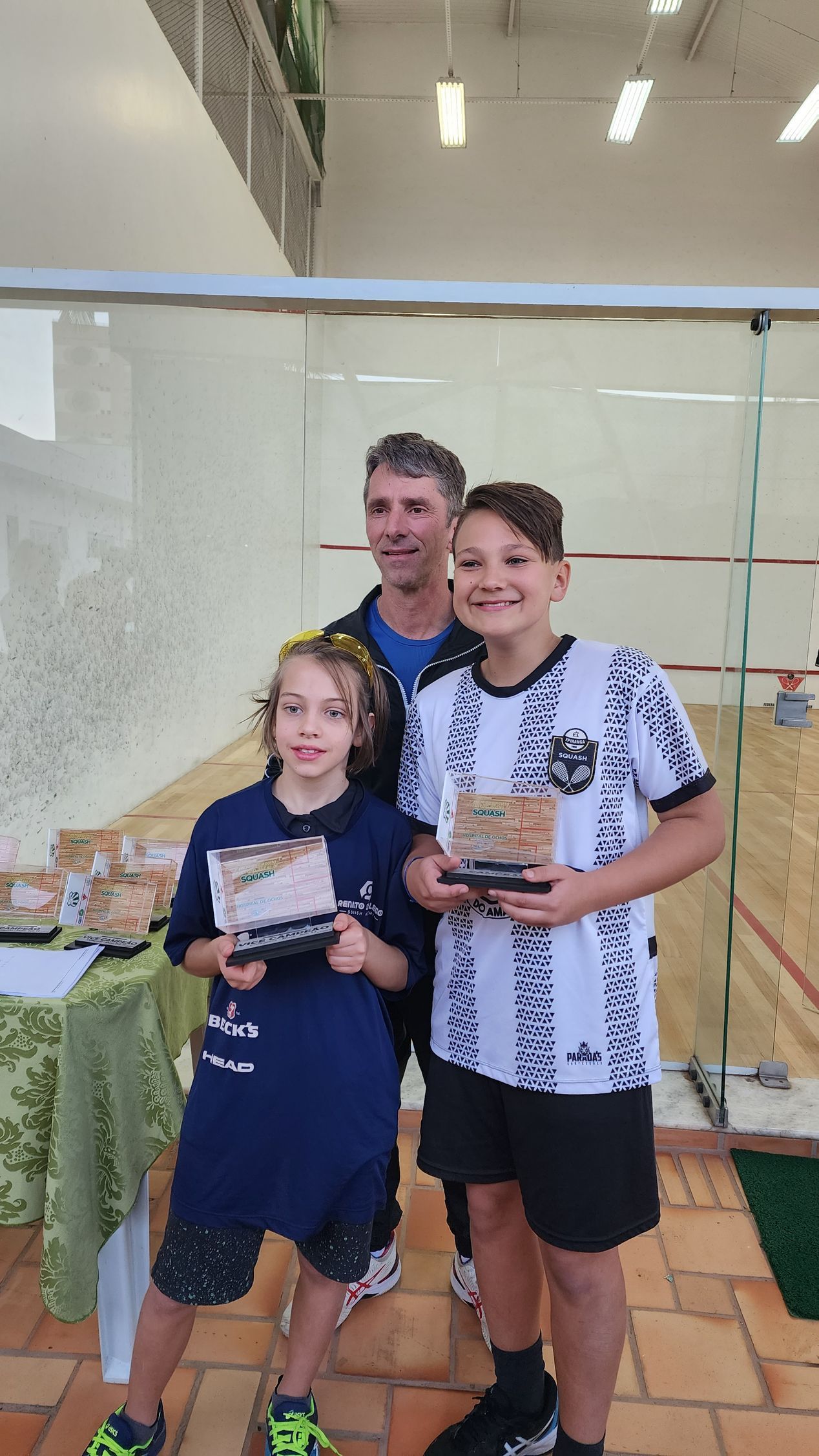 Um homem e dois meninos estão de pé, um ao lado do outro, segurando troféus.