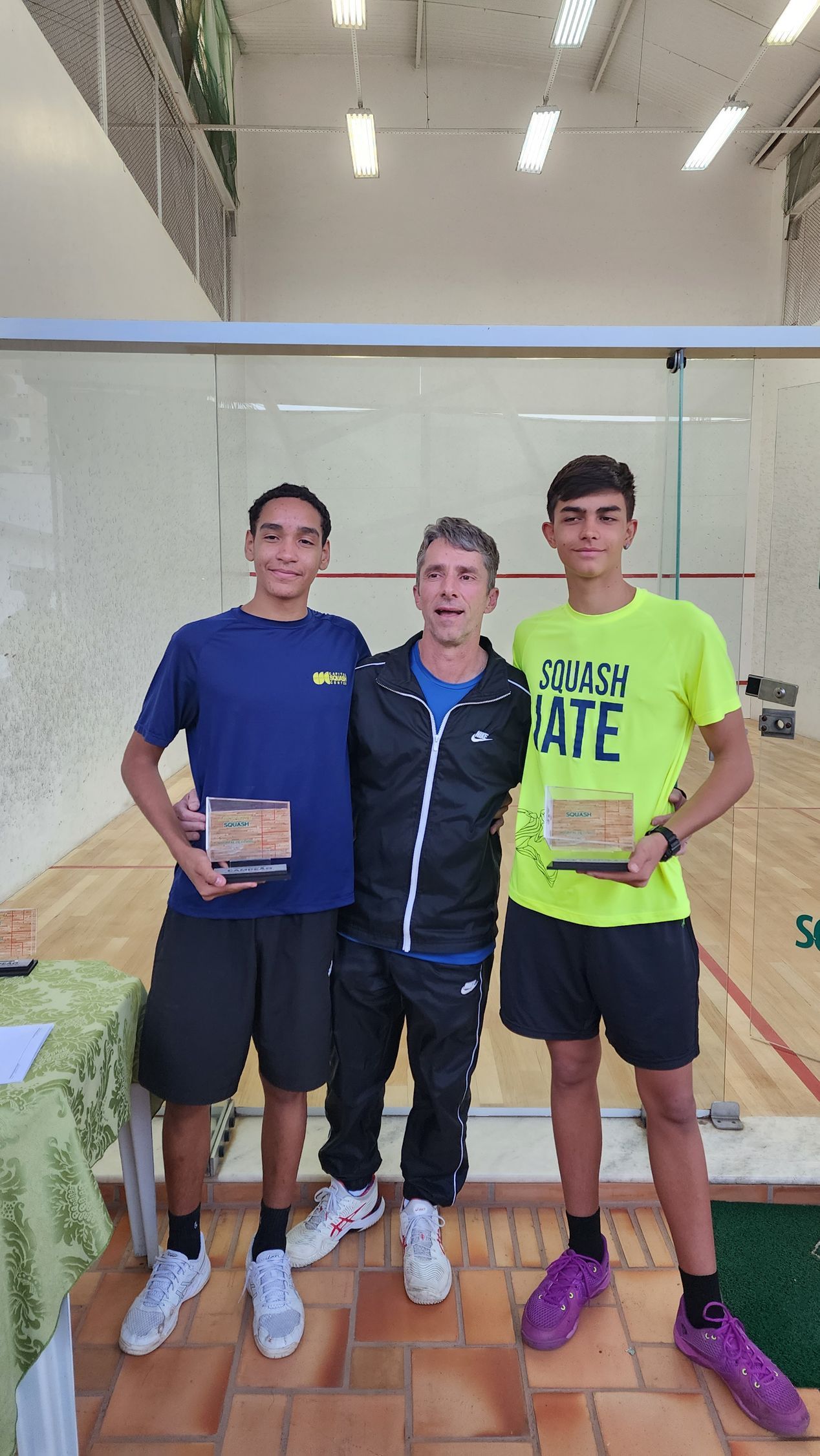 Três jovens estão de pé, um ao lado do outro, em uma quadra de tênis, segurando troféus.