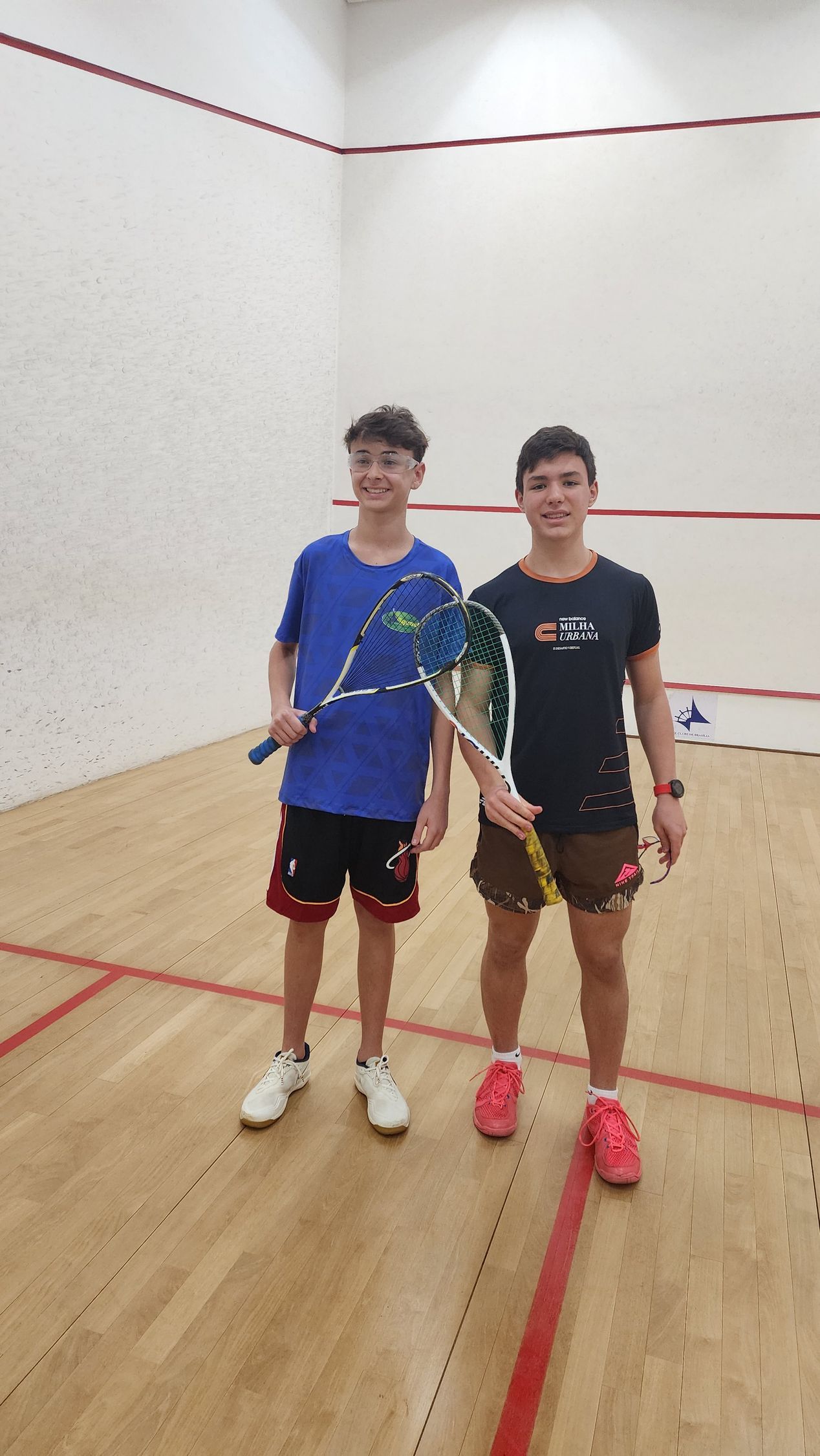 Dois jovens estão em uma quadra de squash segurando raquetes.
