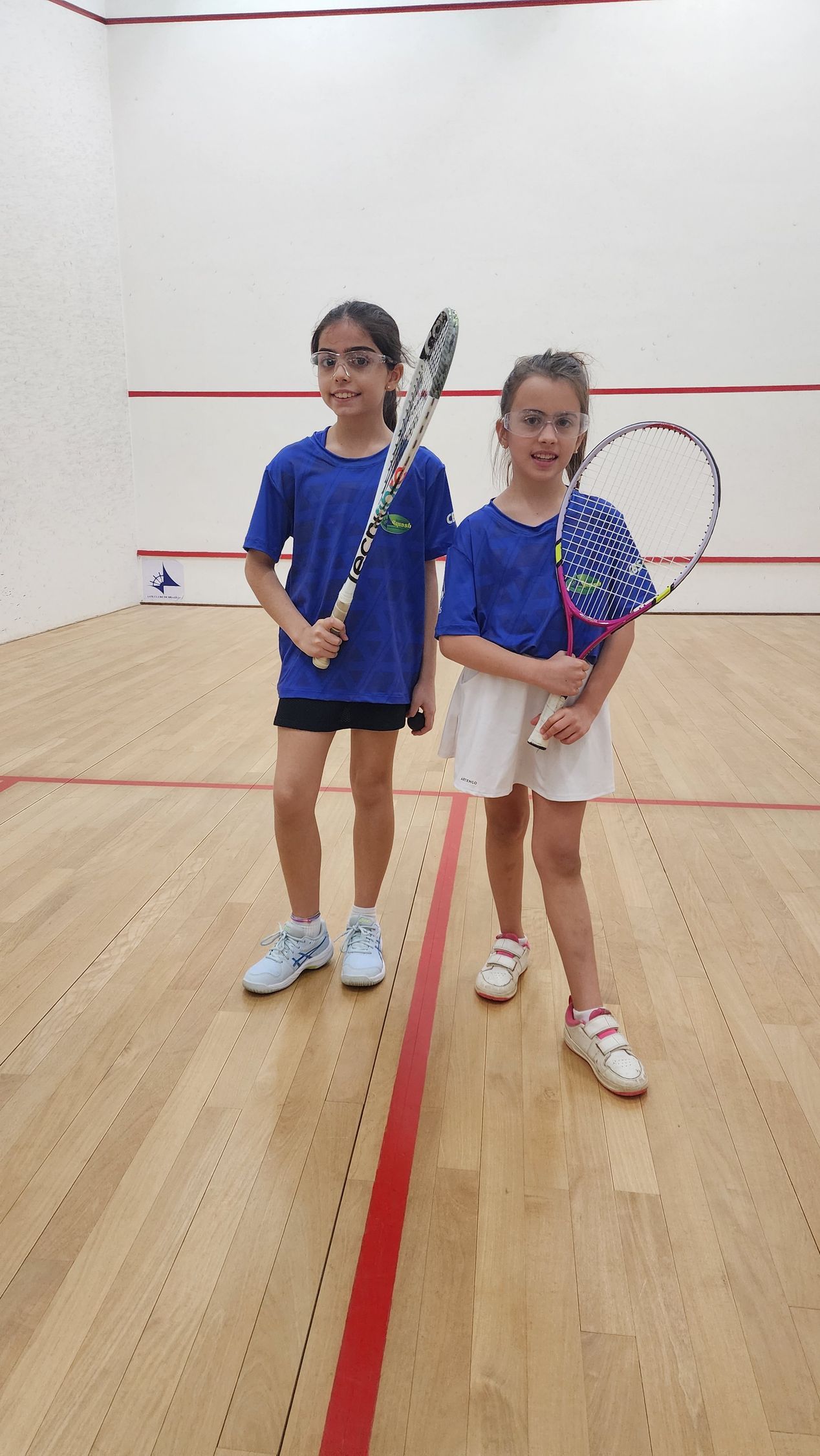 Duas meninas estão em uma quadra de squash segurando raquetes de tênis.