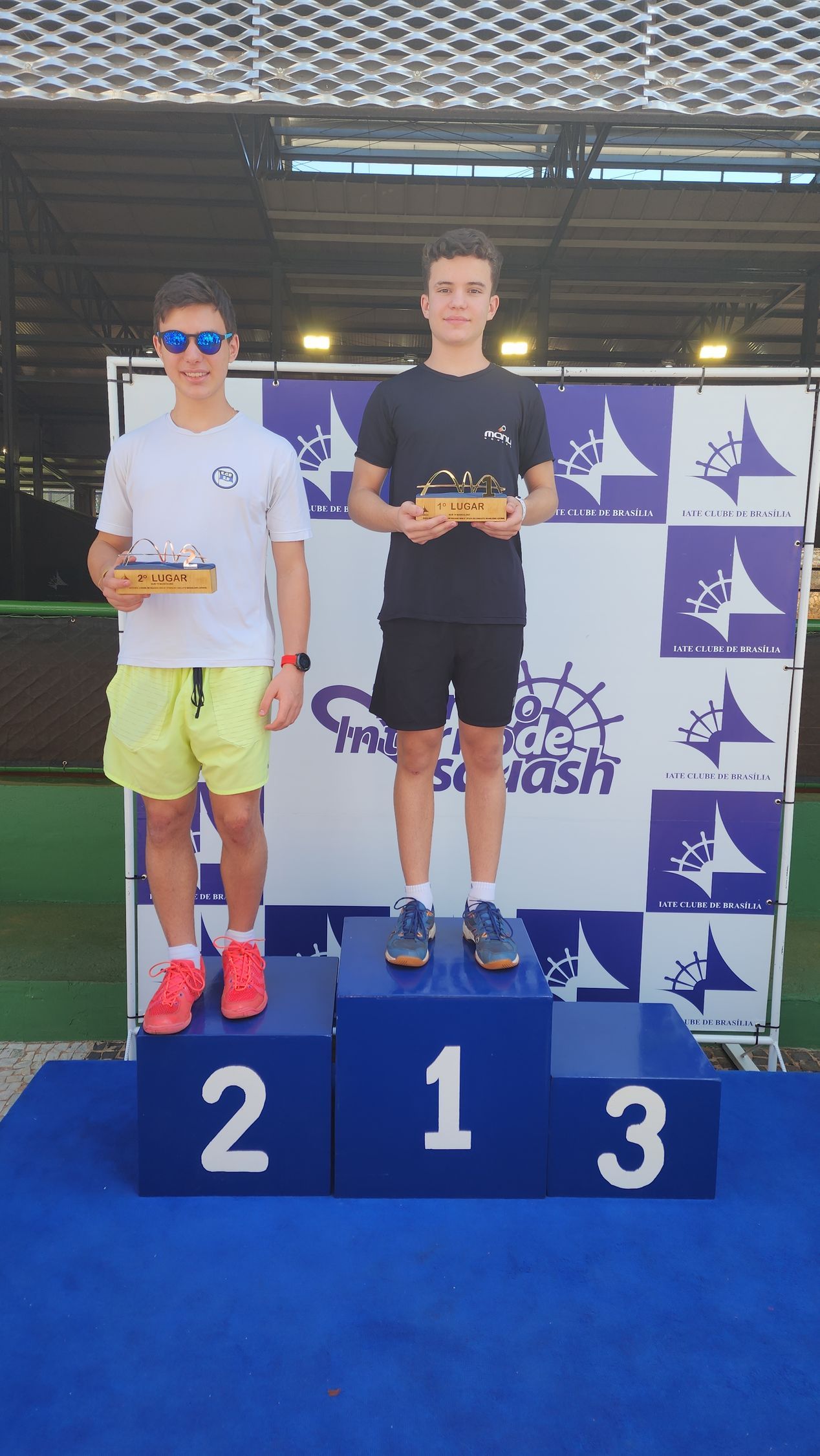 Dois jovens estão em um pódio segurando troféus.