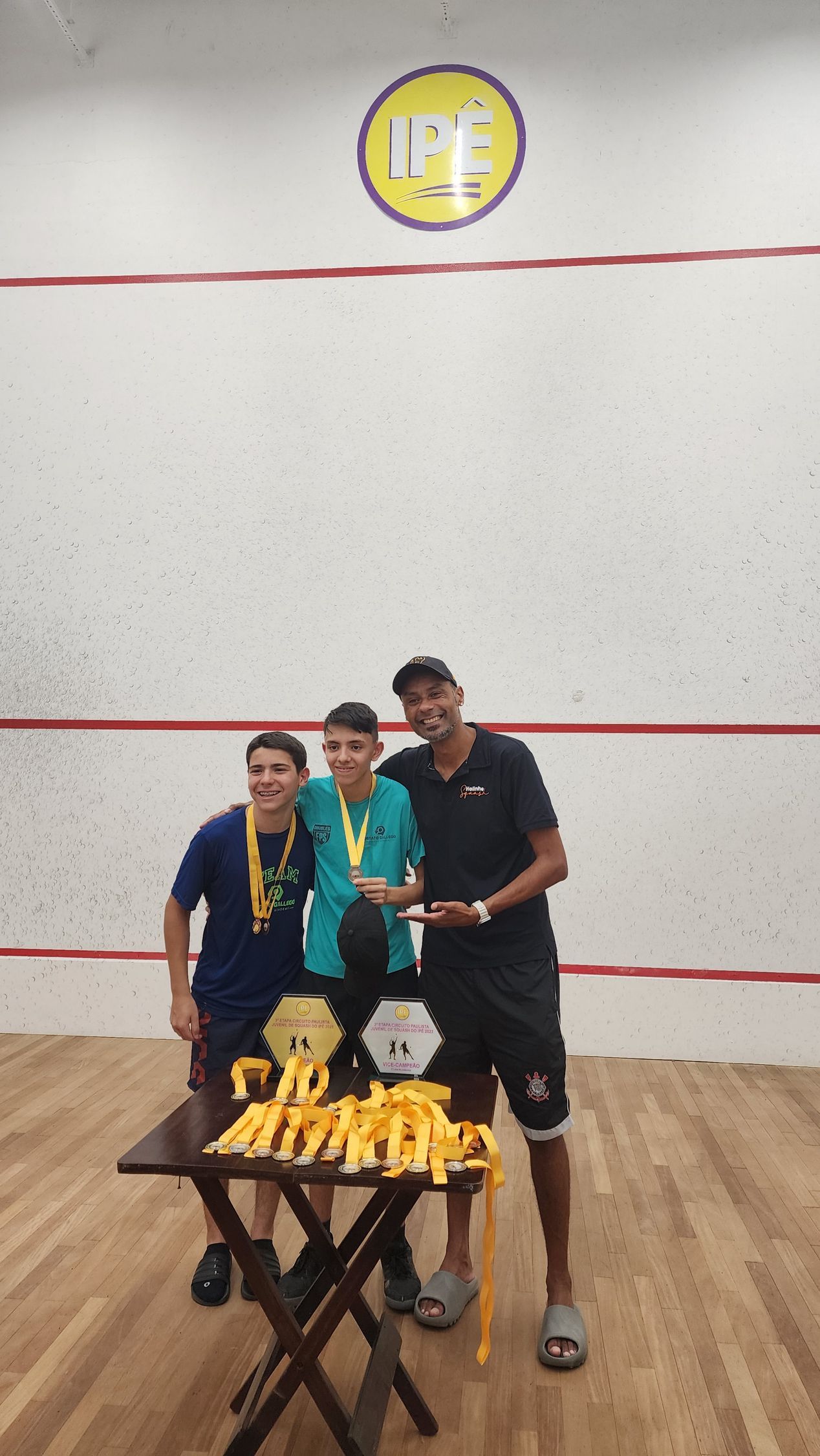 três homens posam para uma foto em frente a uma mesa com troféus.