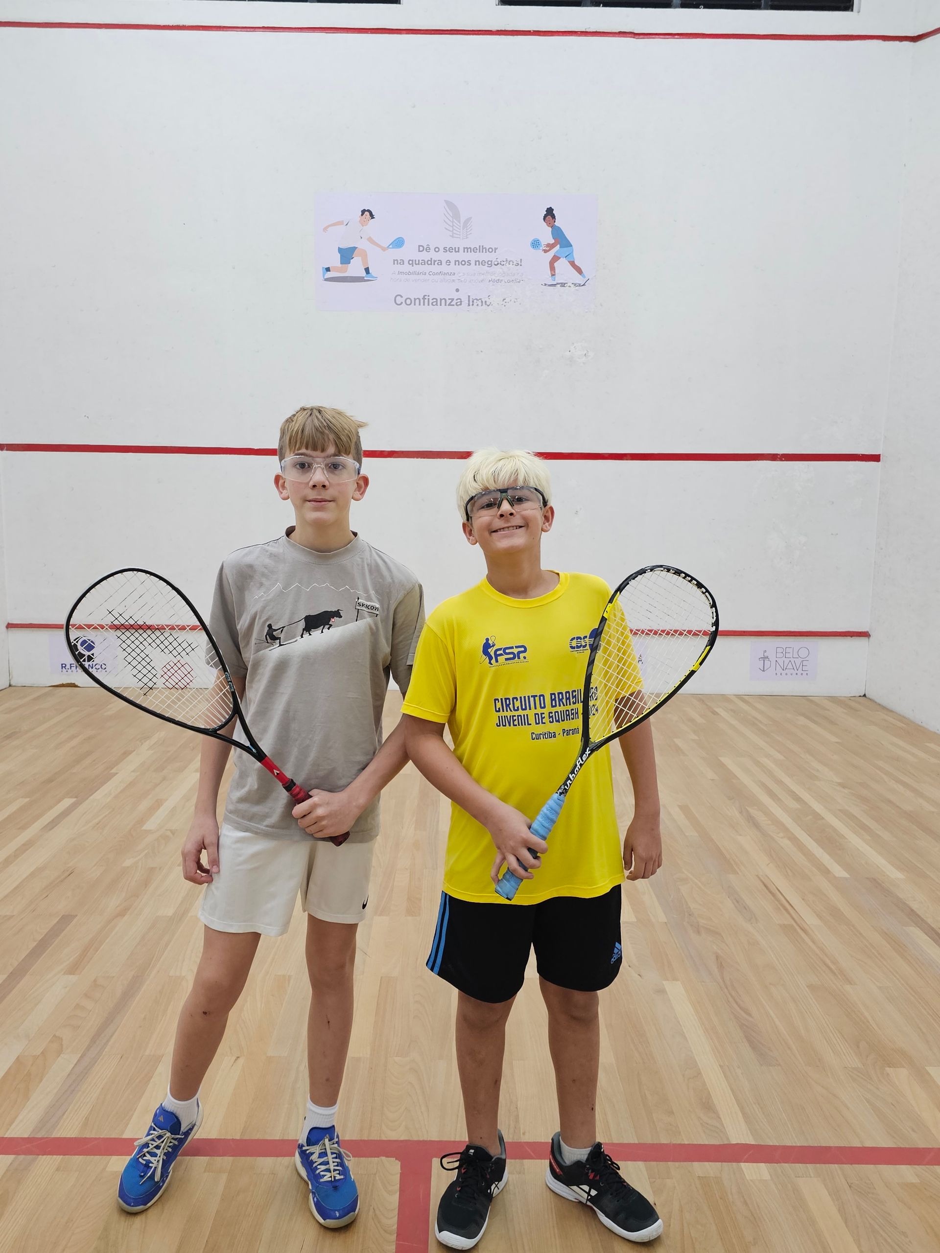 Dois meninos estão em uma quadra de squash segurando raquetes.