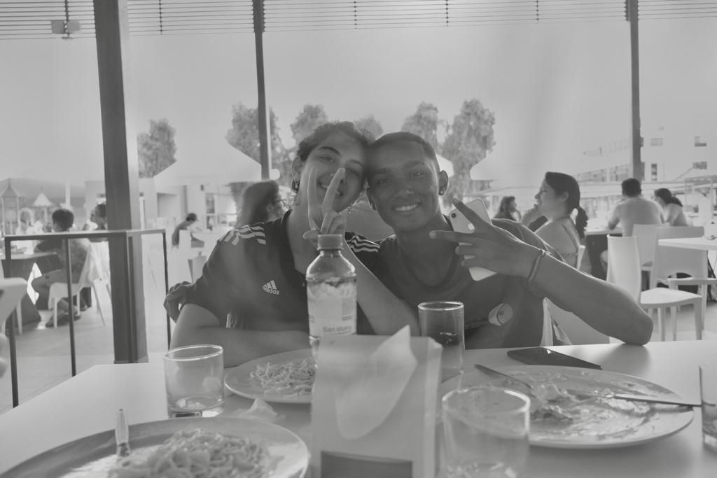 Duas pessoas estão sentadas à mesa com pratos de comida e uma garrafa de água.