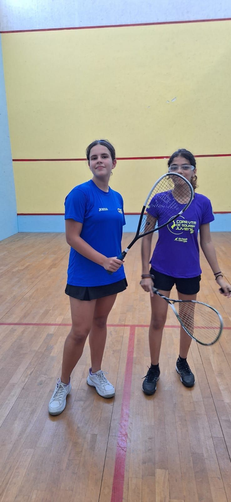 Duas meninas estão de pé, uma ao lado da outra, em uma quadra de squash, segurando raquetes de tênis.