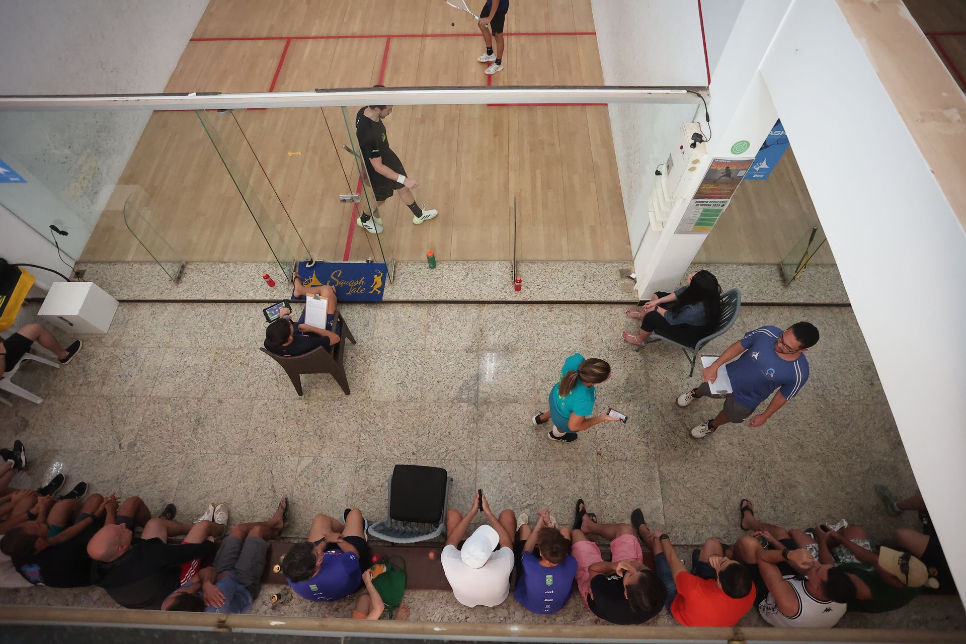 Um grupo de pessoas está sentado no chão em frente a uma quadra de squash.
