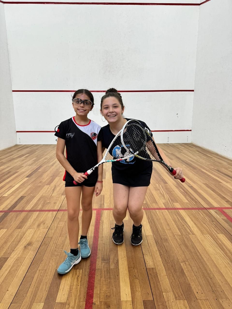 Duas meninas estão em uma quadra de squash segurando raquetes.