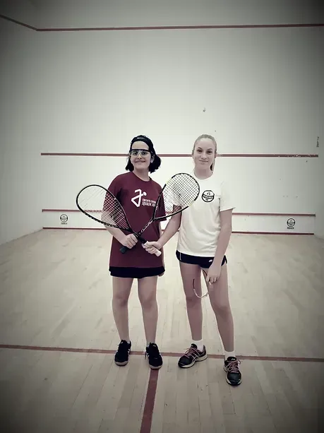 Duas meninas estão em uma quadra de squash segurando raquetes de tênis.