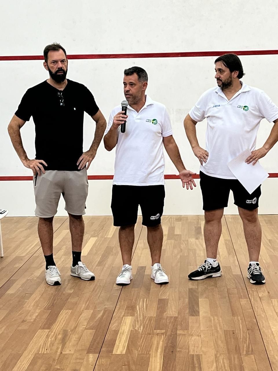 Três homens estão de pé sobre um piso de madeira e um deles está segurando um microfone.