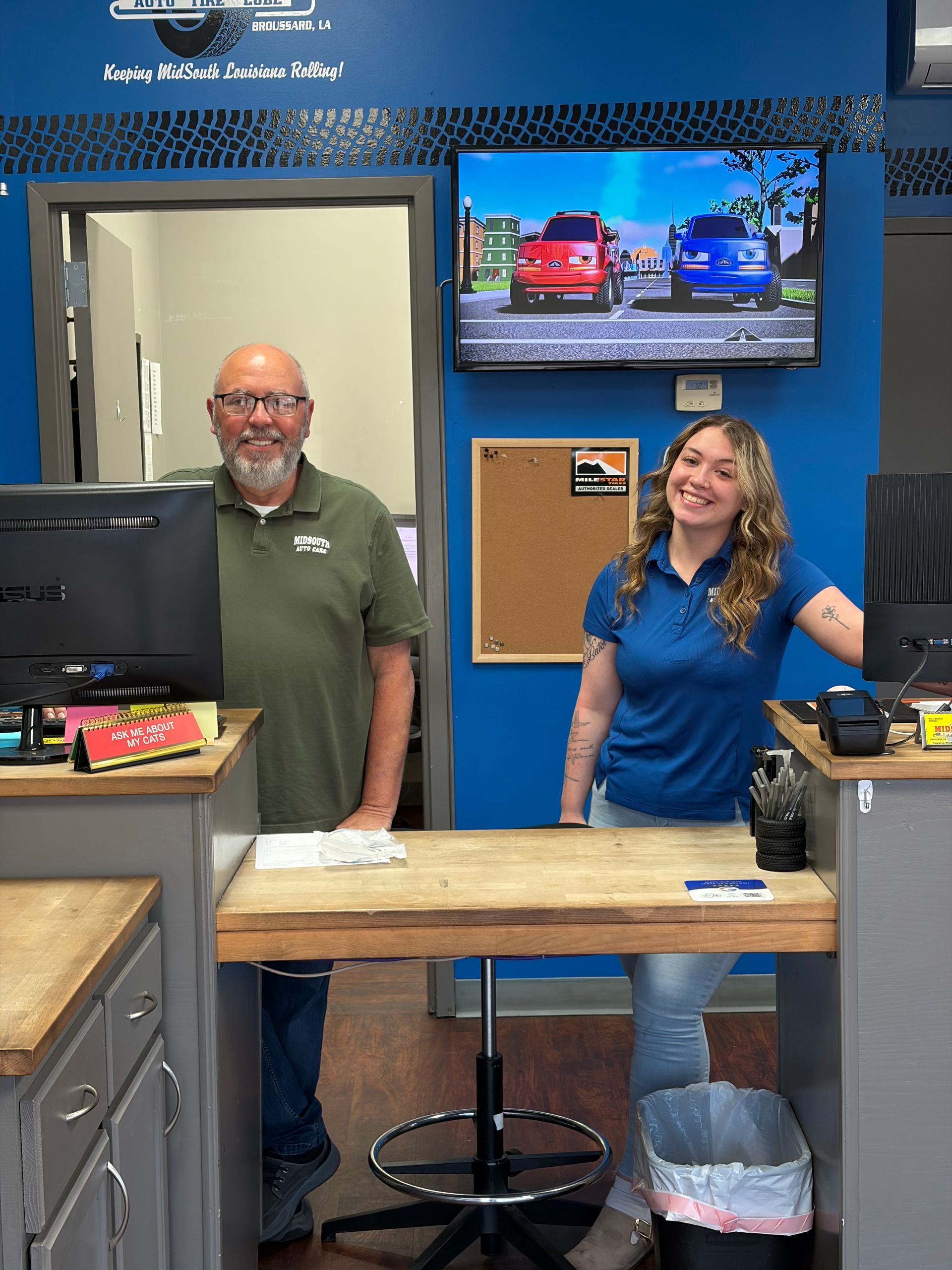 Inside a brake service shop: a man stands behind a desk, two TVs on walls, coffee station, service banner.