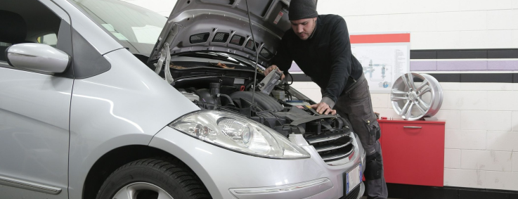 Mechanic working on a silver car with the hood open in a garage.