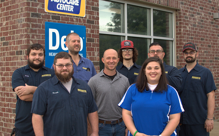 Group of employees standing outside a business, some wearing work shirts, smiling. Brick building and sign visible.
