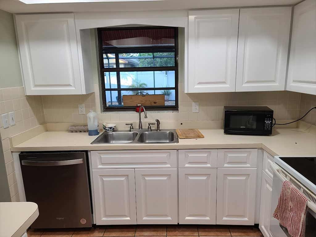 A kitchen with white cabinets , a sink , a microwave , and a window.