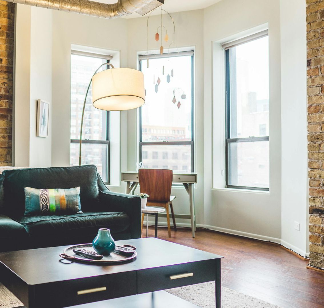Living room with a dark green couch, brick wall, and a desk near windows.
