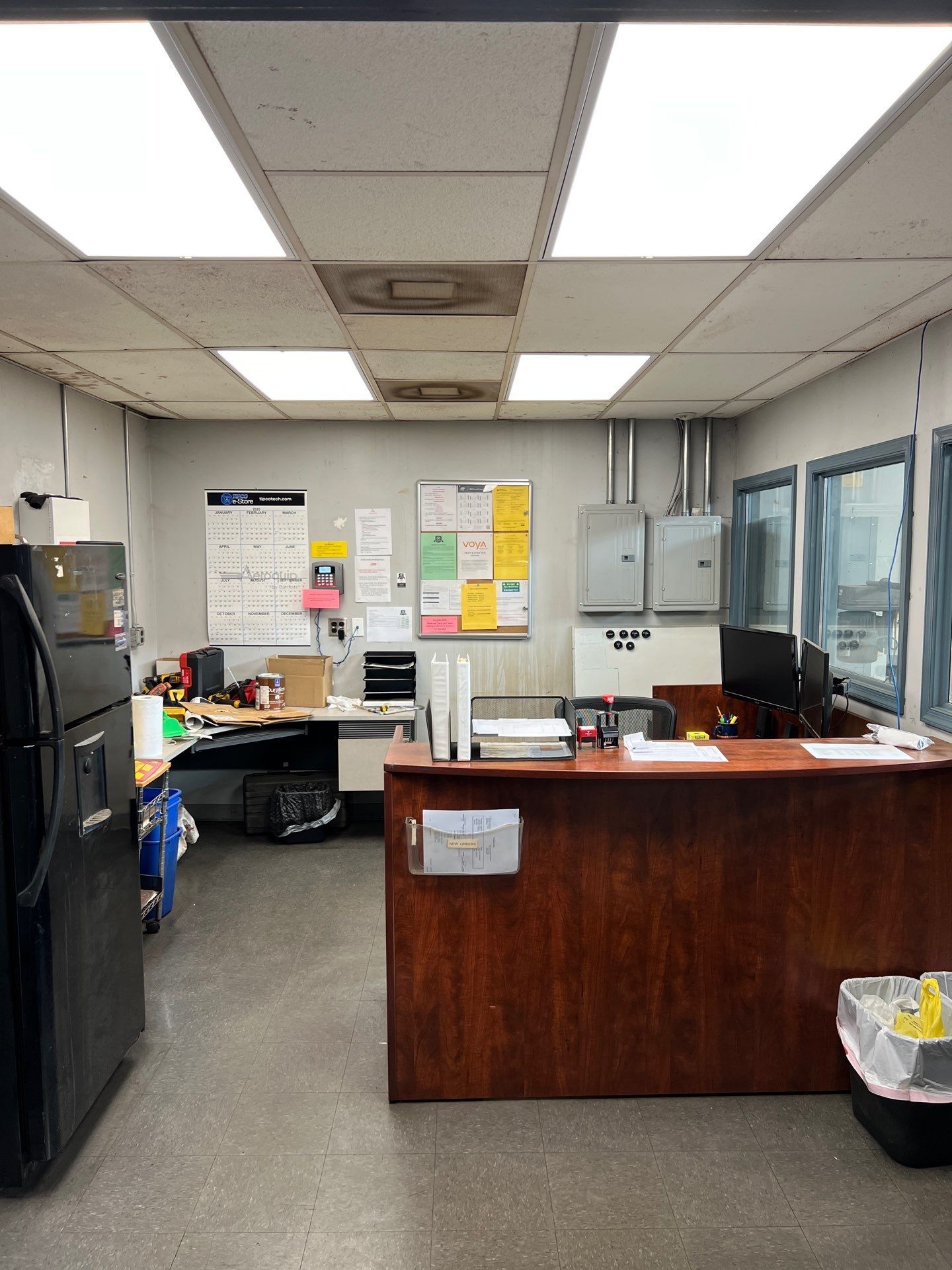 Office space with a desk, refrigerator, and information boards under bright ceiling lights.