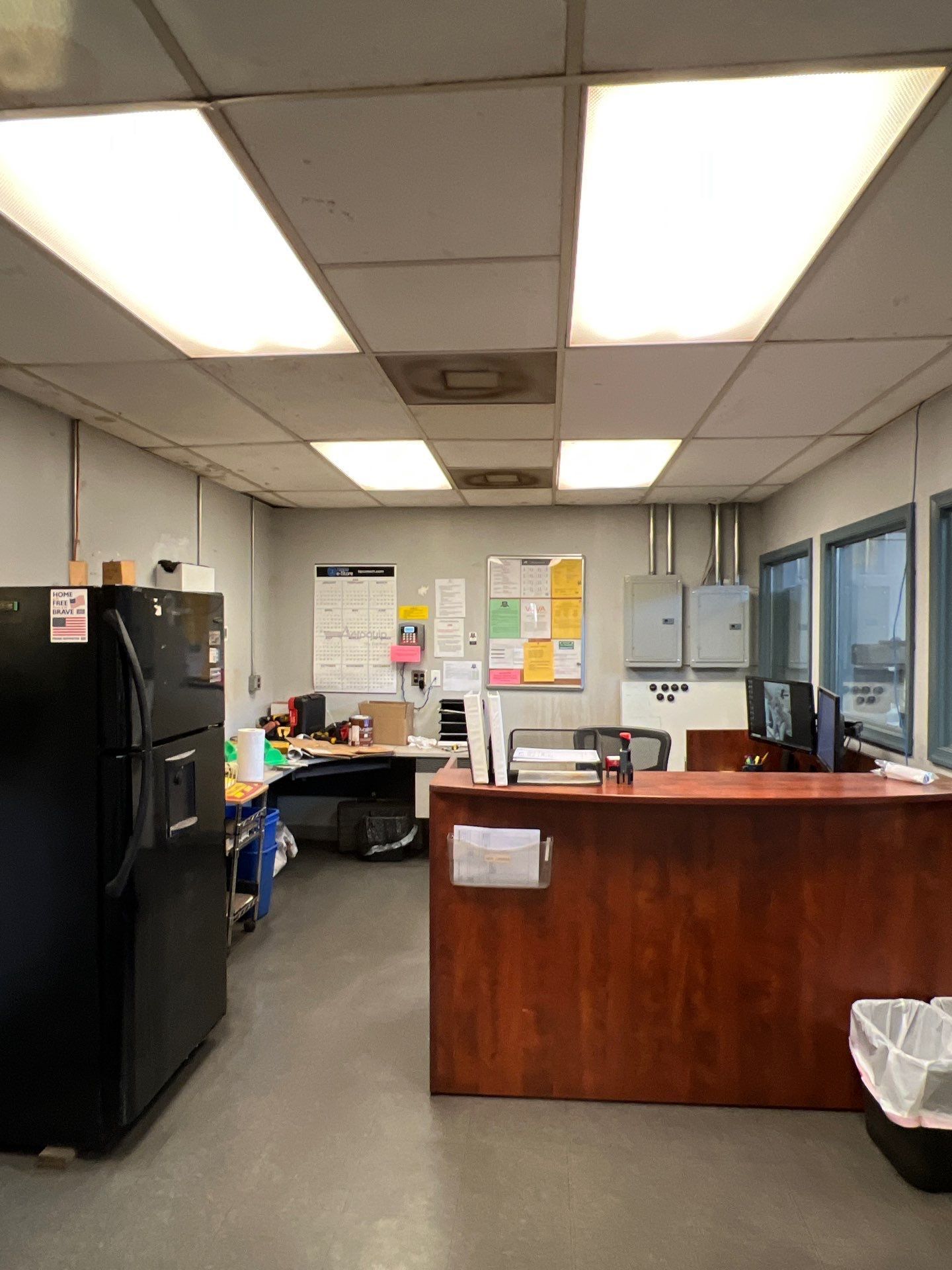Office interior with refrigerator, desk, and reception area, lit by overhead fluorescent lights.