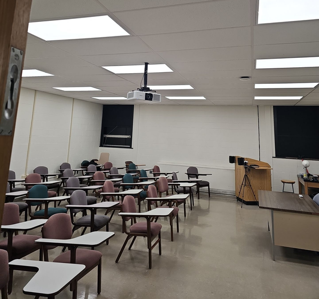 Classroom with rows of student desks, a podium, and a projector.