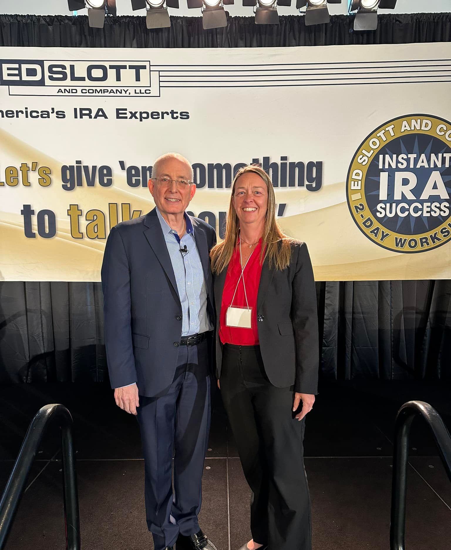 Two people stand on stage in front of a banner. A man and a woman pose with a banner that reads