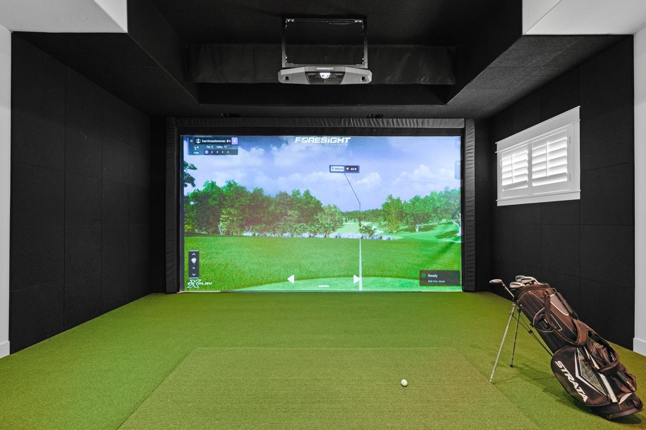 Indoor golf simulator room with a large screen, green turf, and golf bag.