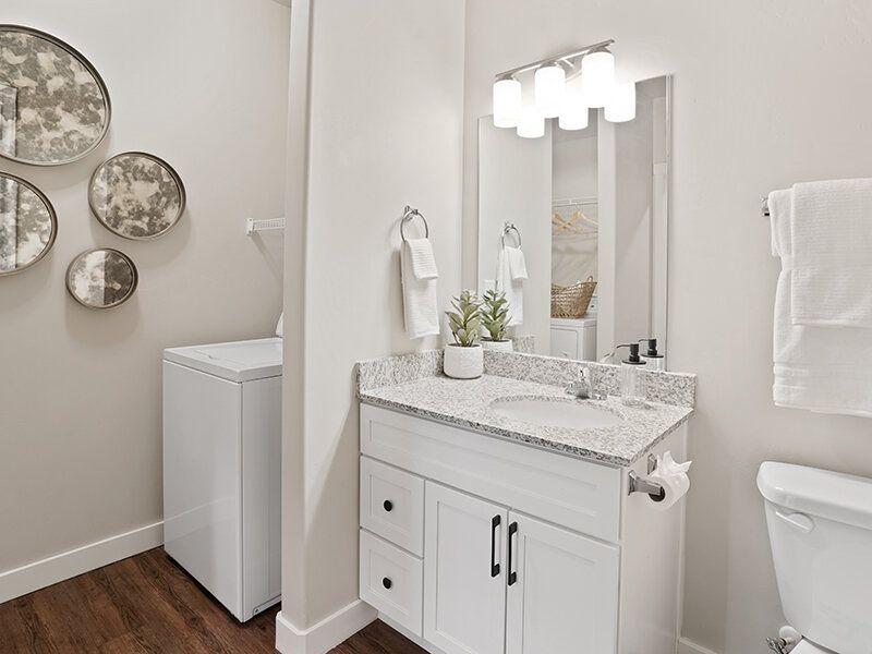 Bright apartment bathroom with a white vanity, granite countertop, mirror, and a nearby washer/dryer.