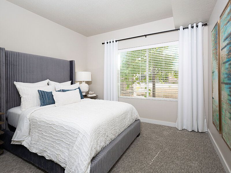 Bedroom in a rental unit with a gray upholstered bed, white bedding, and a window with white curtains.
