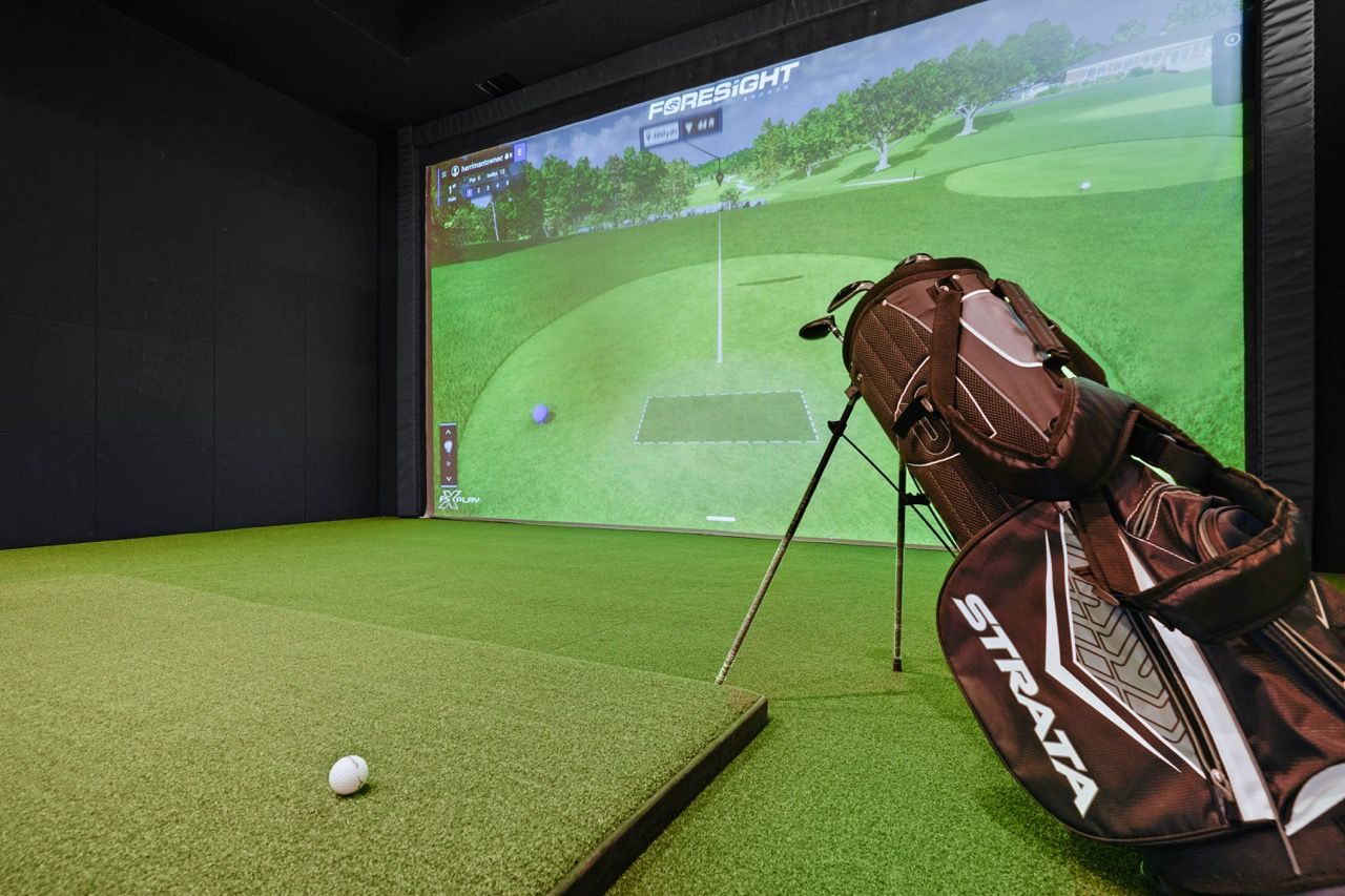 Indoor golf simulator room with turf, a golf bag, and a large screen showing a golf course.