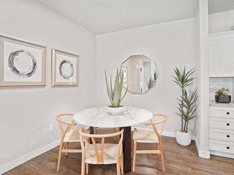 Interior dining area with round marble table, four wooden chairs, plants, and kitchen in the background.