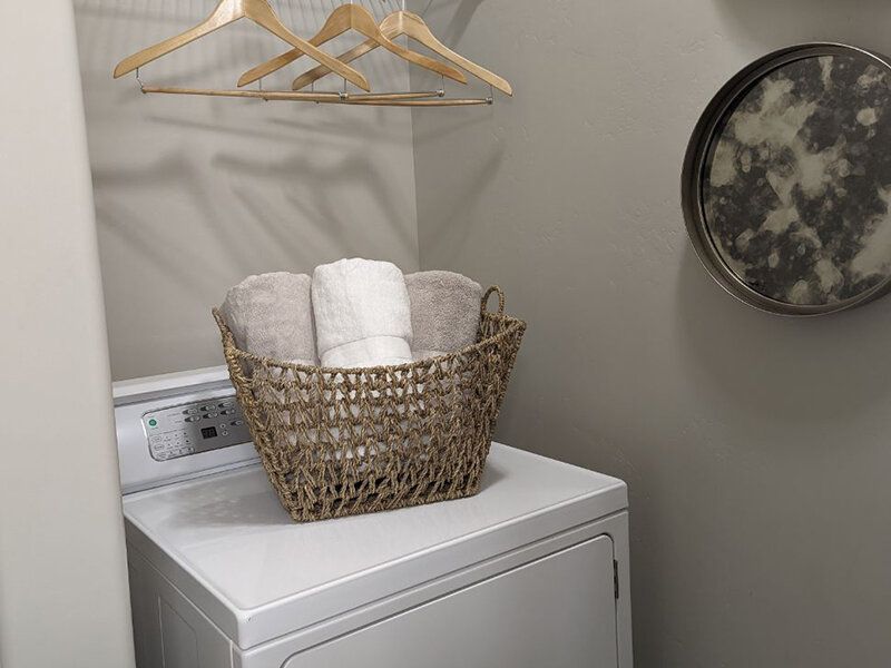 Laundry area with a washing machine, wicker basket of towels, wooden hangers, and a round wall mirror.