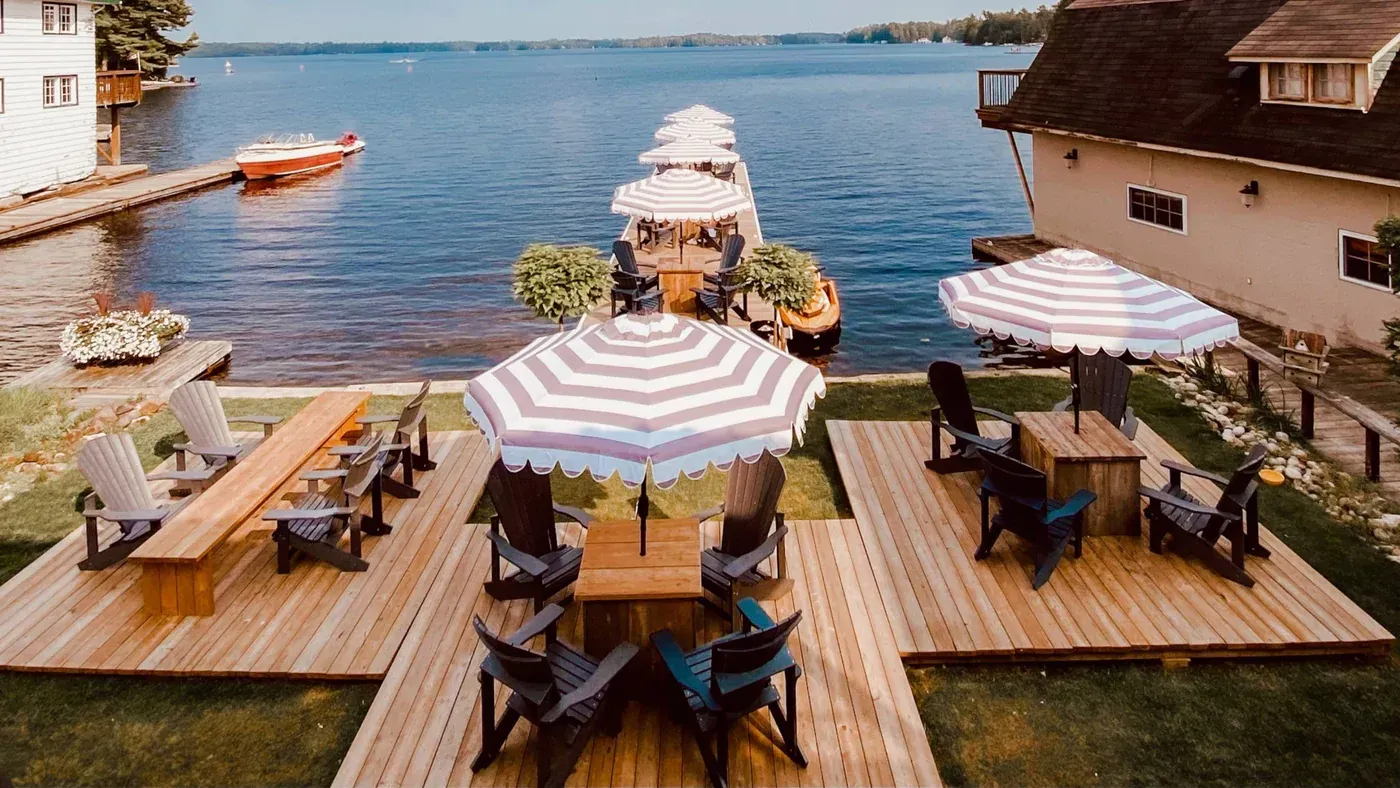 A wooden deck with chairs , tables and umbrellas next to a body of water.