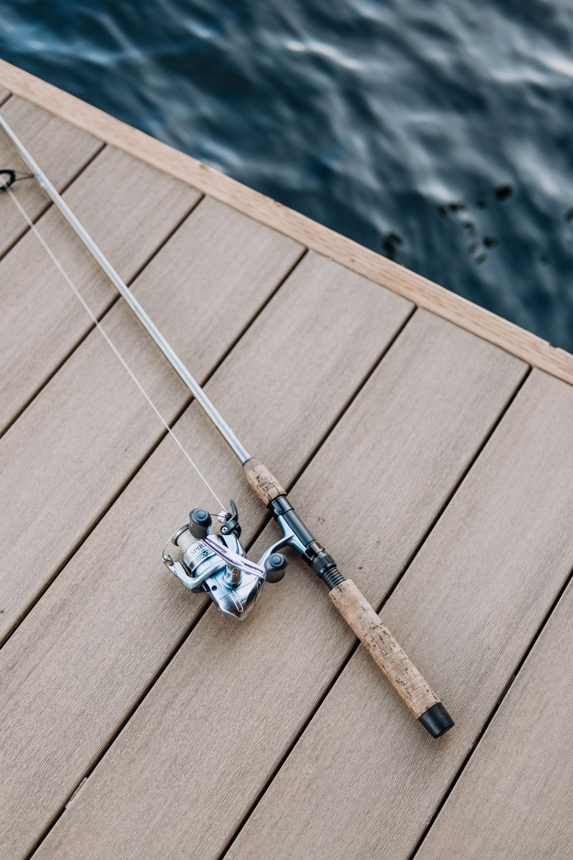 A fishing rod and reel are sitting on a wooden deck next to the water.