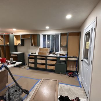 Kitchen renovation in progress: dark green cabinets installed, light wood drawers, tools scattered, doorway on right.
