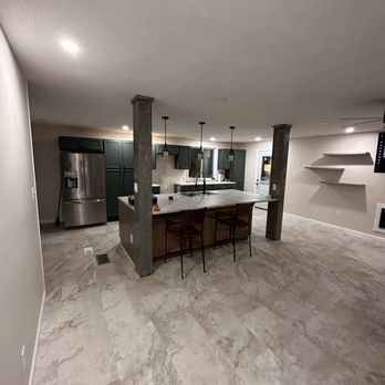 Open kitchen with dark green cabinets, island with stools, stainless steel refrigerator, and tiled floor.
