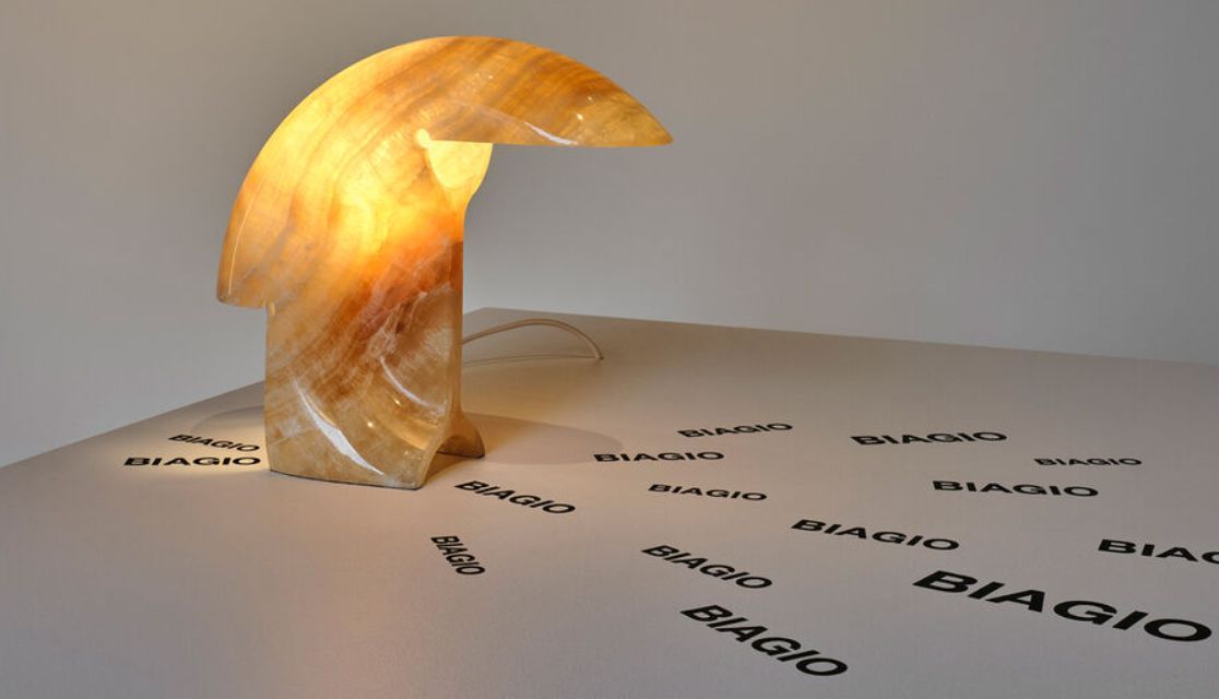 Scultura a mezzaluna color ambra traslucida su una superficie bianca con ombre di testo nere sparse.