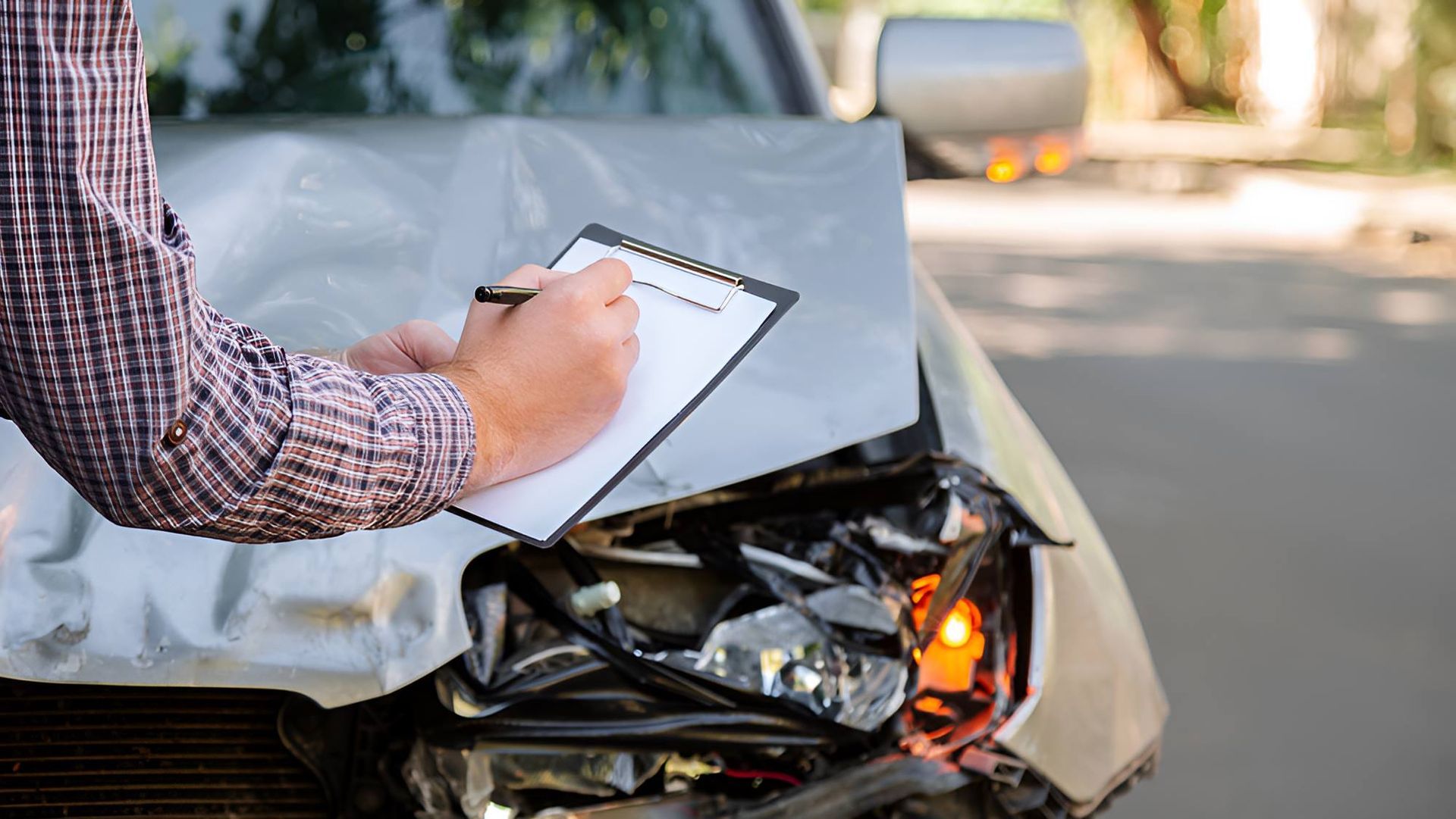 A Man is Writing on a Clipboard in Front of a Damaged Car — Optimus 1 Insurance Brokers in Westcourt, QLD