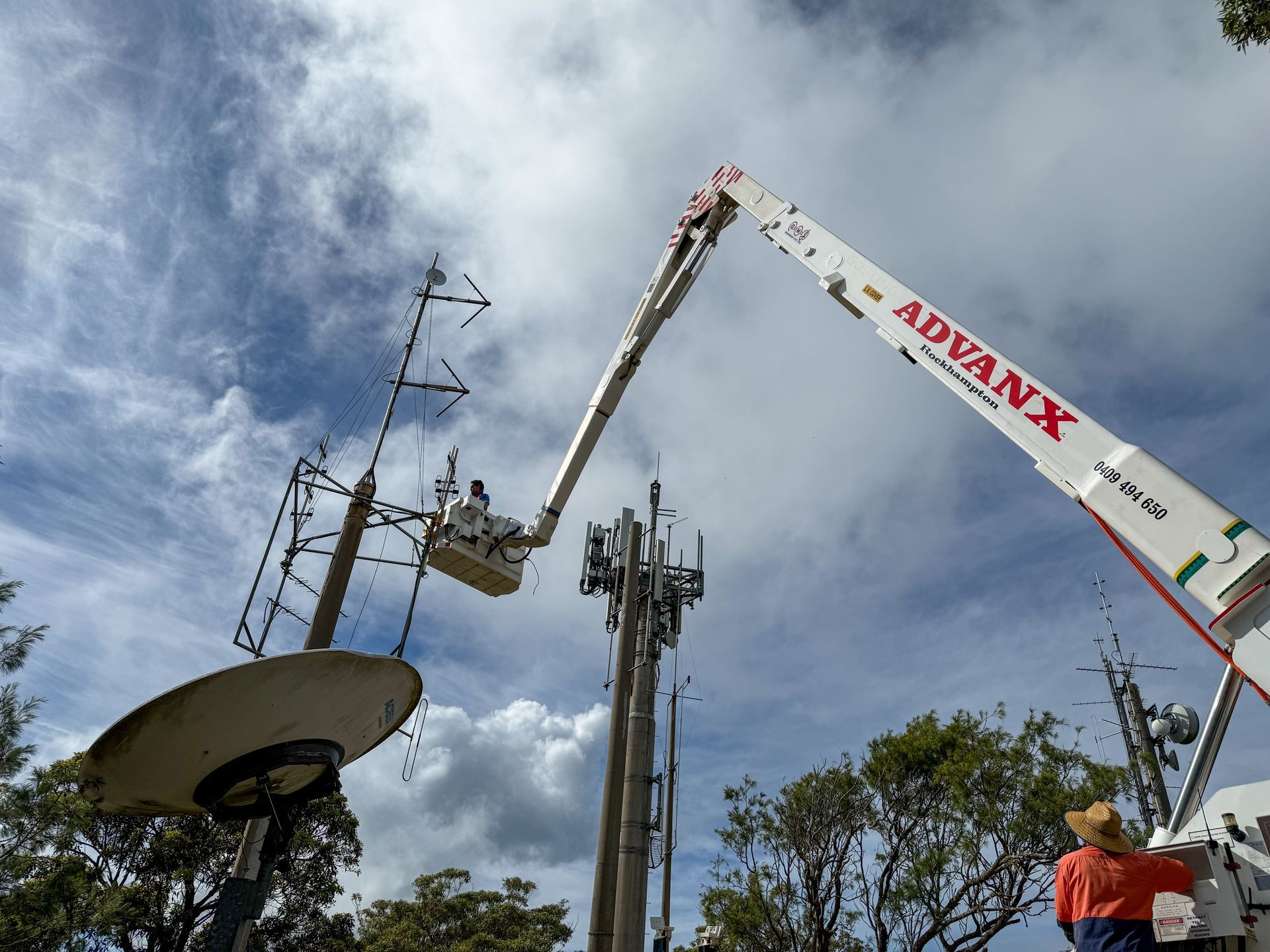 A worker in a lift basket is working on a cell tower antenna, under a partly cloudy sky.