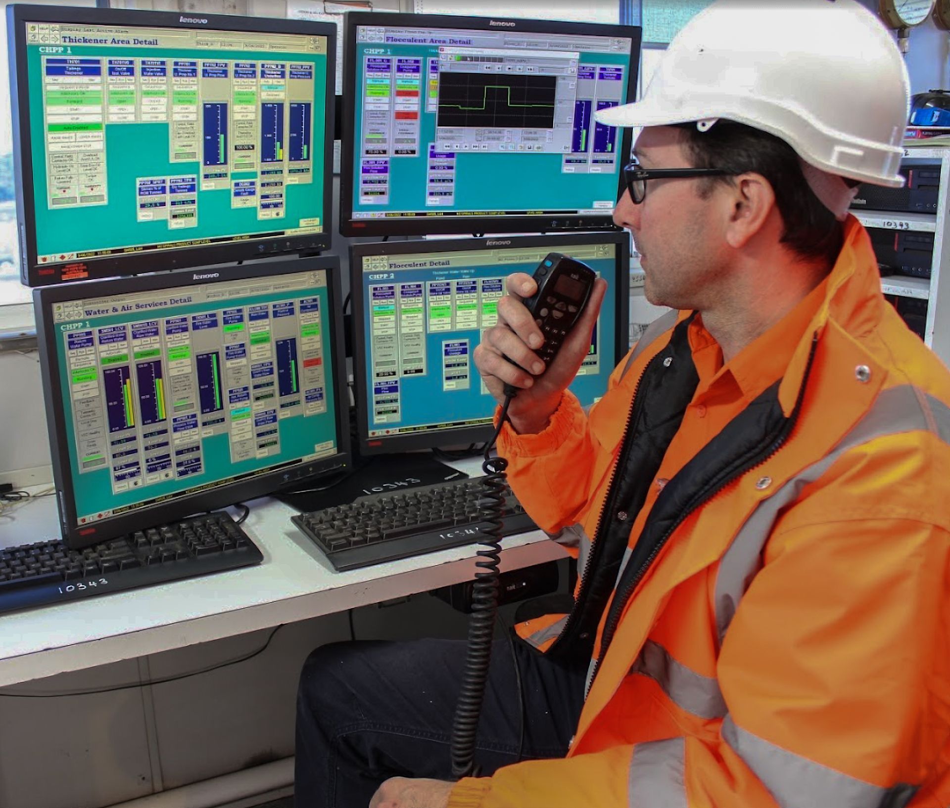 Man in safety vest and hard hat using a handheld radio, monitoring computer screens.
