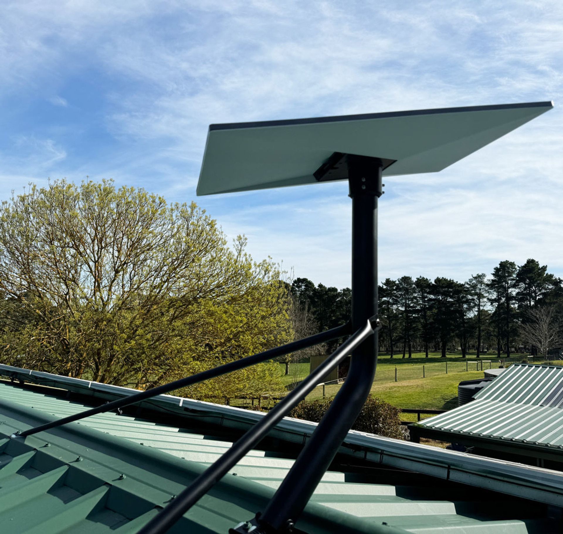 Black satellite dish mounted on a green roof against a blue sky, trees, and field.