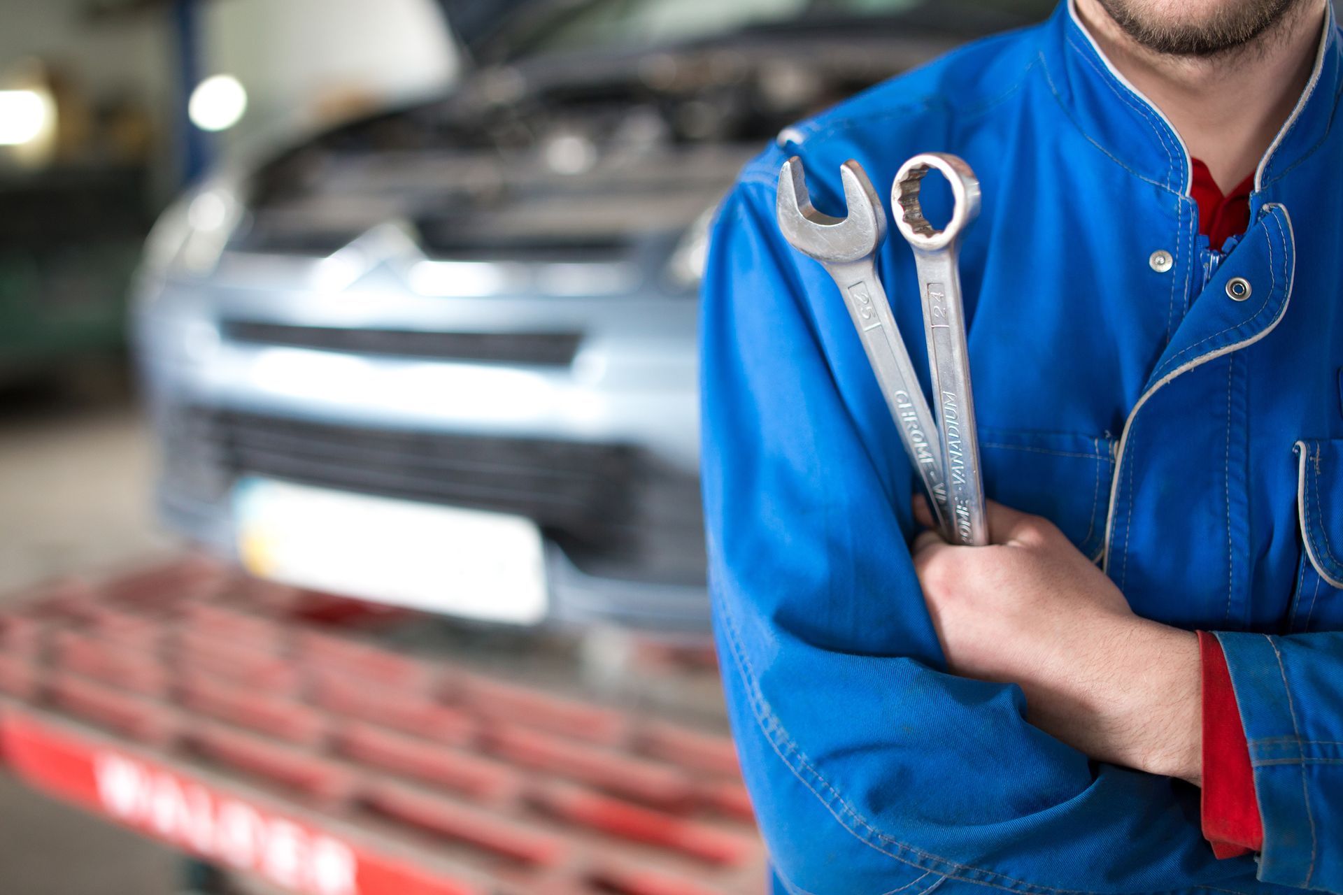 A man in a blue uniform is holding two wrenches in front of a car.