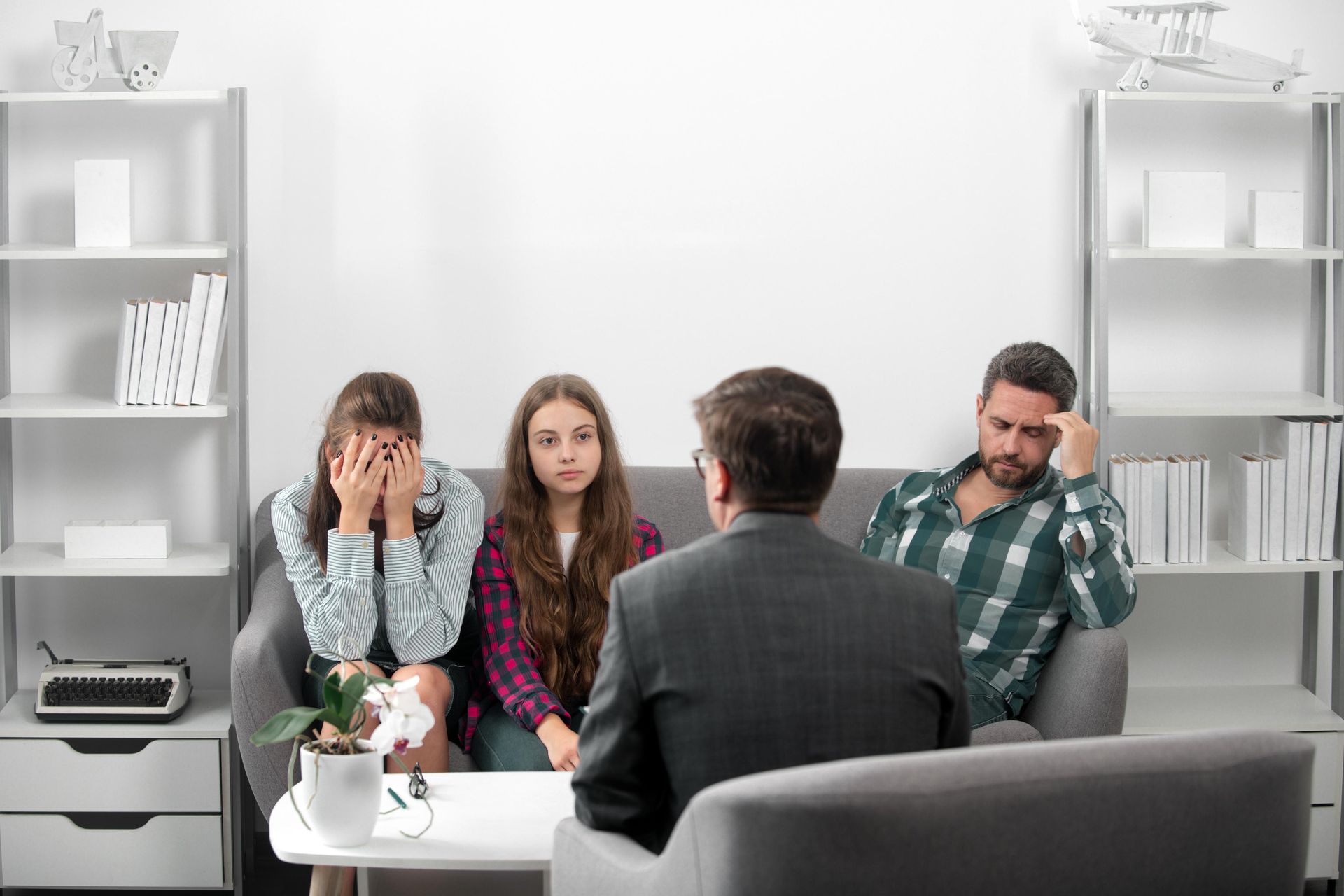 Family in therapy session; parents and children on couch, facing therapist. Sad expressions and concerned gestures.