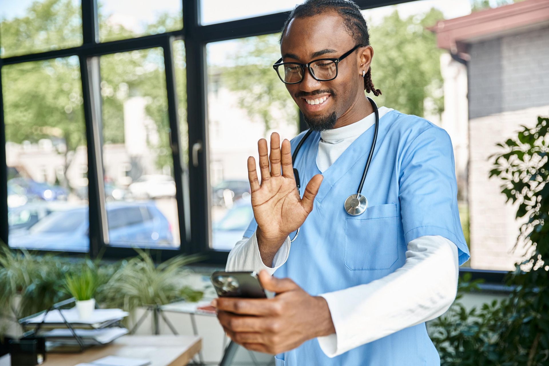 Doctor in blue scrubs waves at phone screen, smiling. Stethoscope around neck, near window.