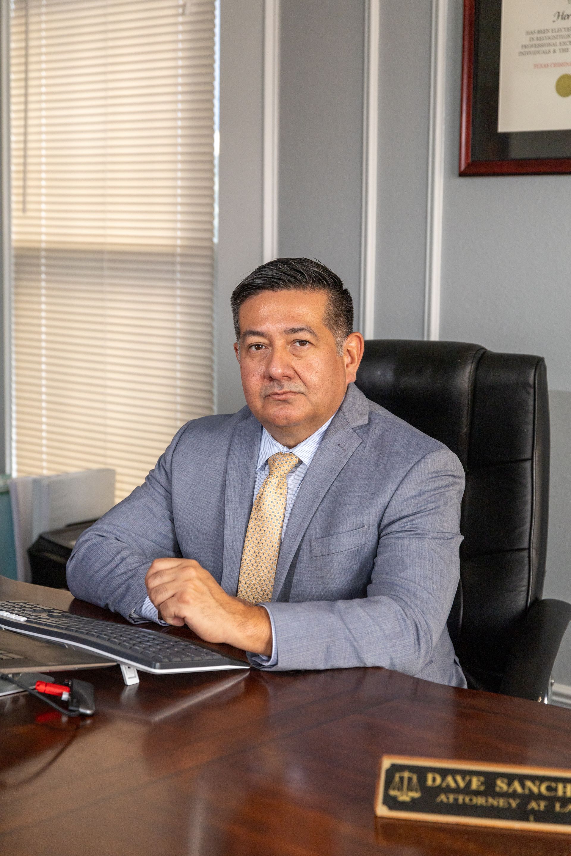 Man in suit, attorney at desk in office. Brown hair, neutral expression, window blinds, and legal document in background.