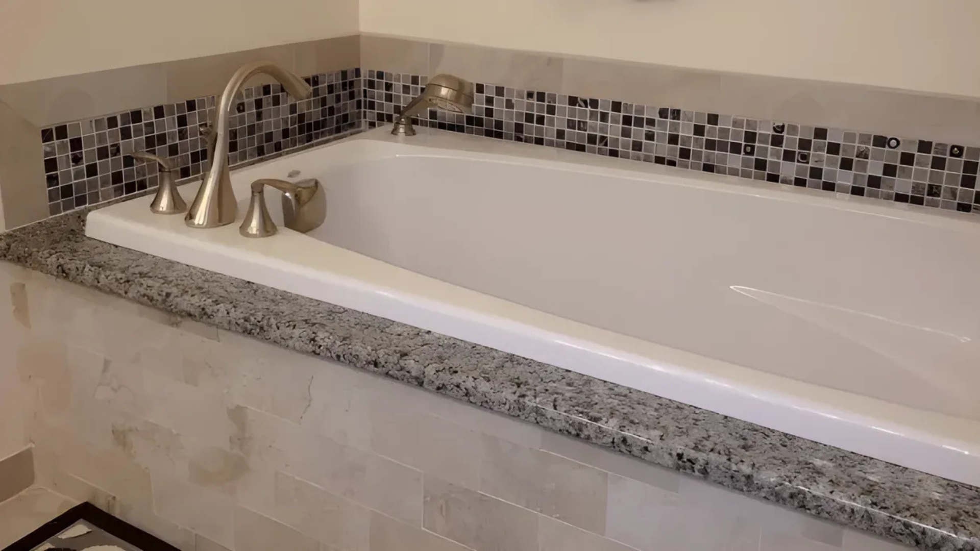 Bathtub with granite countertop, tile backsplash, and brushed nickel fixtures.
