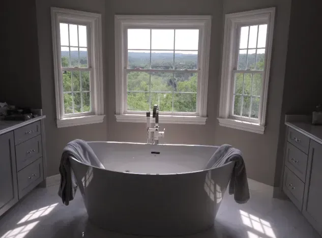 Freestanding white bathtub centered beneath three windows with a view of a green forest.