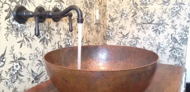 Copper sink with water flowing from a dark faucet, set against a patterned wallpaper.