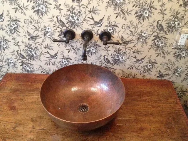 Copper bowl sink on wooden table with wall-mounted faucet against floral wallpaper.