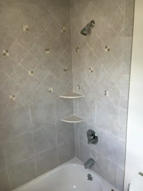 Corner shower with gray tile, two shelves, and a chrome shower head and fixtures.